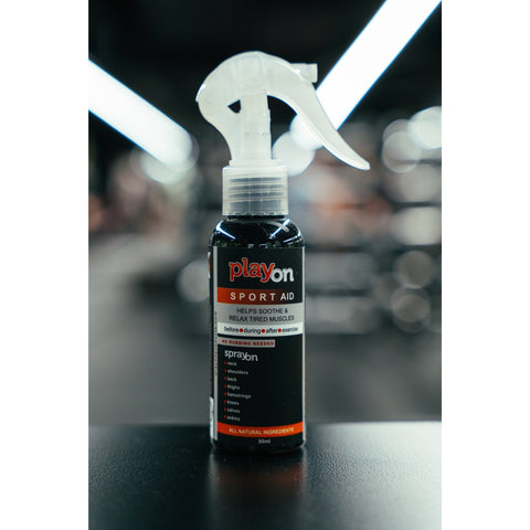 A bottle of Playon Sport Aid 80ml spray with a white trigger sprayer and black label, set on a dark surface with a blurred gym background.